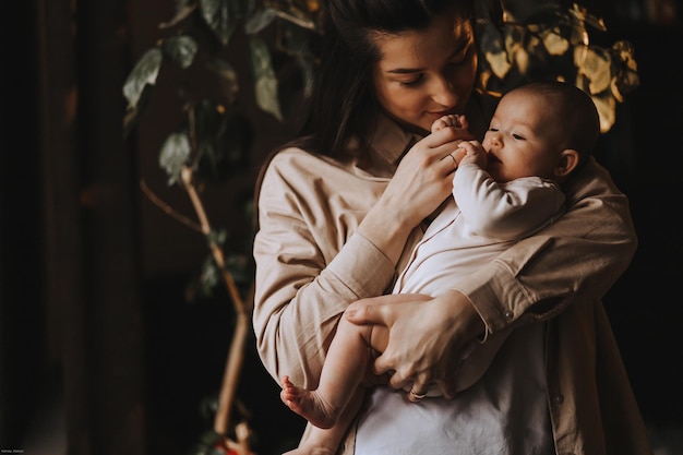 Jeune maman brune et son nouveau-né dans ses bras photo à la maison dans une pièce sombre près de la fenêtre