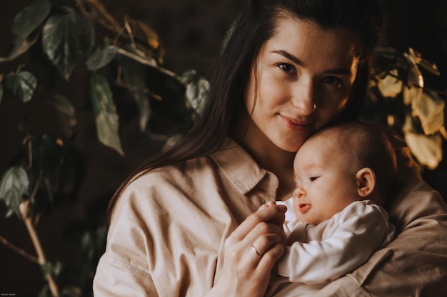 Jeune maman brune et son nouveau-né dans ses bras photo à la maison dans une pièce sombre près de la fenêtre