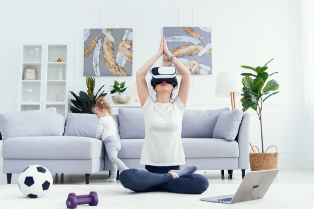 Jeune maman méditant en position de yoga lotus à l'aide de lunettes ar pendant que sa fille regarde des dessins animés sur