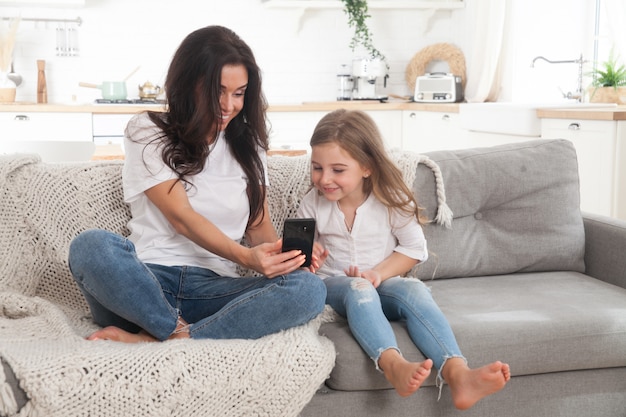 Jeune maman et petite fille jouant à des jeux en ligne à la maison à l'aide d'un smartphone