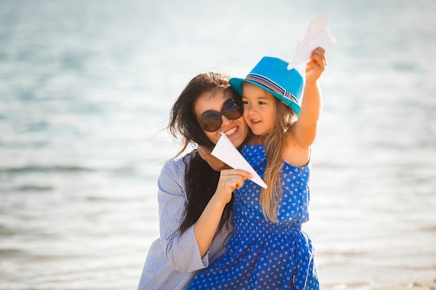 Jeune maman et sa jolie fille au bord de la mer lançant des avions en papier dans l'air et riant. Famille joyeuse à la plage