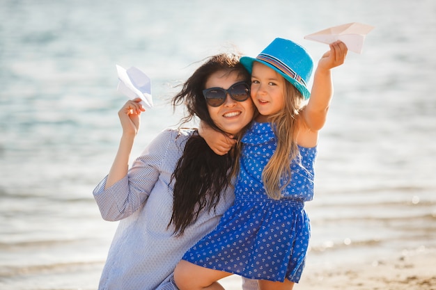 Jeune maman et sa jolie fille au bord de la mer lançant des avions en papier dans l'air et riant. Famille joyeuse à la plage