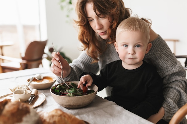 Jeune maman séduisante aux cheveux rouges en pull tricoté assis à la table, nourrissant pensivement son petit fils qui rêveusement