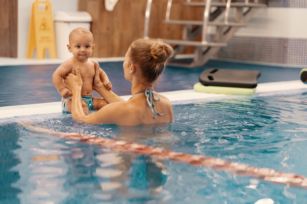 Jeune maman et son bébé bénéficiant d'une leçon de natation bébé dans la piscine. Enfant s'amusant dans l'eau avec maman