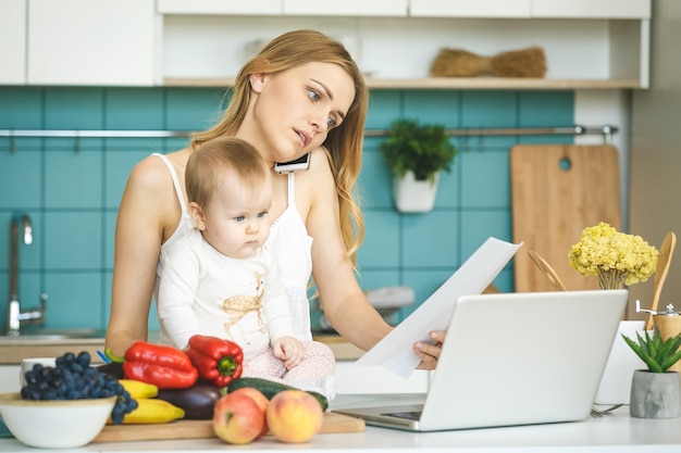 Jeune maman travaille à la maison. Cuisiner et jouer avec sa petite fille dans une cuisine moderne. En regardant un ordinateur portable.