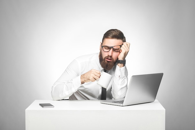 Un jeune manager fatigué et irresponsable en chemise blanche et cravate noire est assis au bureau et essaie de ne pas dormir sur le travail, boit une tasse de café, tenant la tête avec la main. Studio shot, isolé, intérieur