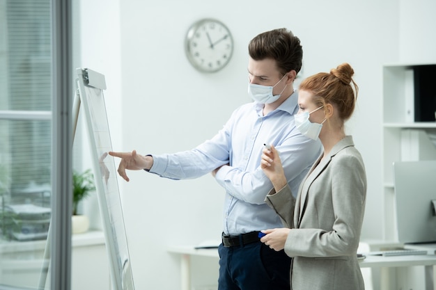 Jeune manager masculin portant un masque de protection pointant sur un tableau blanc tout en expliquant les données financières à sa collègue féminine avec surligneur