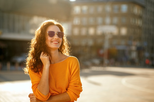 Jeune mannequin gai aux cheveux bouclés dans des lunettes de soleil portant une tenue tendance et marchant dans la rue ensoleillée. Espace pour le texte