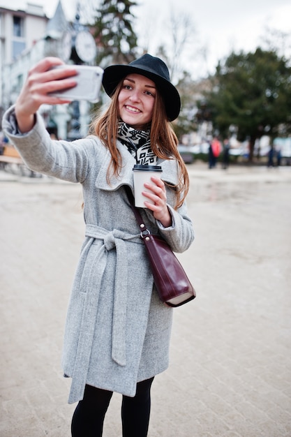 Jeune Mannequin Portant Un Manteau Gris Et Un Chapeau Noir Avec Un Sac à Main En Cuir Sur Les épaules Et Une Tasse De Café En Plastique