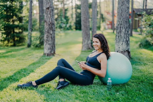 Une jeune mannequin sportive détendue prend une pause après des exercices d'aérobic avec un ballon de fitness assis sur l'herbe verte utilise des conversations sur téléphone portable en ligne écoute de la musique dans des écouteurs a un corps athlétique