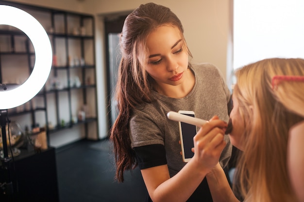 Jeune maquilleuse appliquant le fard à joues sur les joues de la femme blonde dans la salle de beauté. Regard professionnel sur le client.