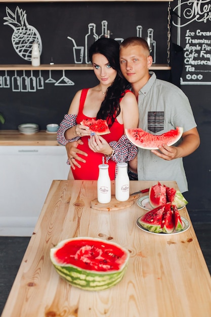 Jeune mari étreignant sa belle femme enceinte en haut rouge et chemise Ils tiennent une pastèque en tranches à une table en bois dans une cuisine moderne