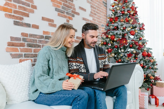 Le jeune mari et la femme de la famille choisissent ensemble des cadeaux de Noël dans une boutique en ligne, assis à la maison près de l'arbre de Noël avec un ordinateur portable