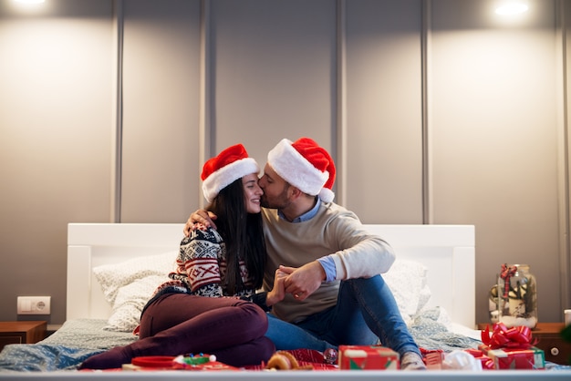 Jeune mari et femme souriant assis sur le lit tout en étreignant et en embrassant avec des chapeaux de Noël et des cadeaux.