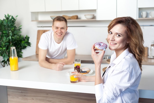 Jeune mari heureux dans la cuisine s'occupe de sa femme. Femme mangeant un beignet et buvant du jus. Lovestory, couple amoureux, gens heureux, futurs parents regardant la caméra et sourire à pleines dents