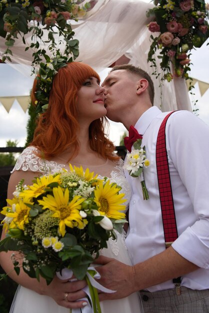 Photo le jeune marié embrasse la jeune mariée aux cheveux roux sur la joue lors de sa cérémonie de mariage en plein air.