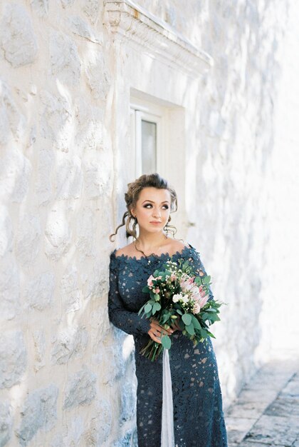 La jeune mariée dans une robe bleue de dentelle s'est penchée contre le mur d'une maison en pierre