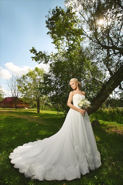 Jeune mariée à la mode, belle fille blonde modèle avec une coiffure de mariage élégante, en robe de dentelle blanche avec bouquet de fleurs dans ses mains posant à l'extérieur au coucher du soleil