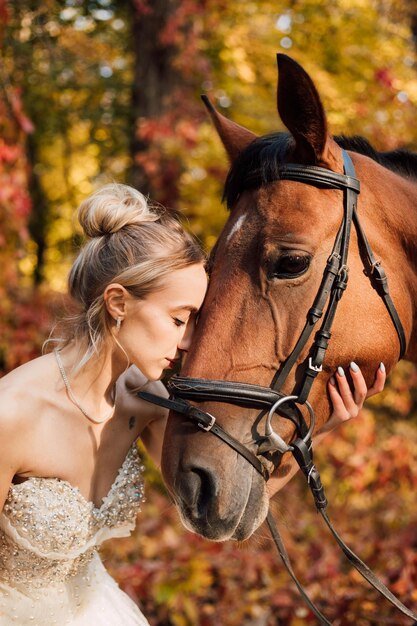 Jeune mariée tendre dans une robe blanche luxuriante posant dans un parc d'automne avec un cheval par une journée ensoleillée