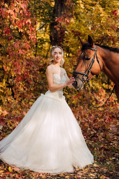 Jeune mariée tendre dans une robe blanche luxuriante posant dans un parc d'automne avec un cheval par une journée ensoleillée