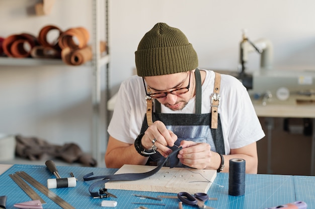 Jeune maroquinier créatif se penchant sur la table tout en cousant un nouvel article en cuir noir sur planche de bois en atelier