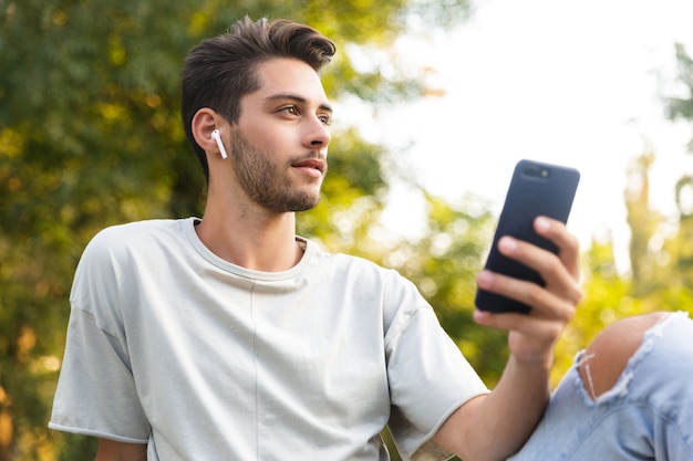 Jeune mec assis dans le parc à l'extérieur à l'aide de téléphone mobile, écouter de la musique avec des écouteurs