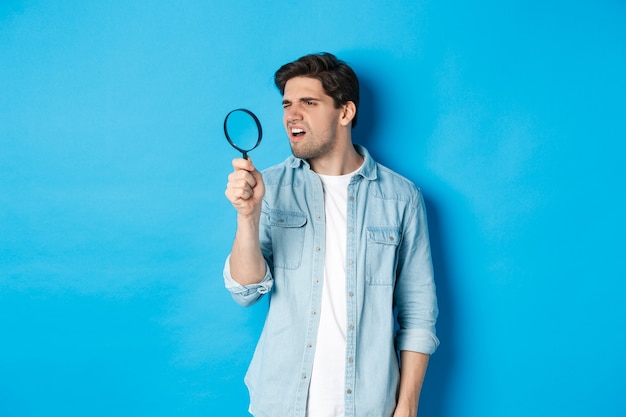 Jeune mec attentionné à la loupe, lisant quelque chose de minuscule, debout sur un mur bleu