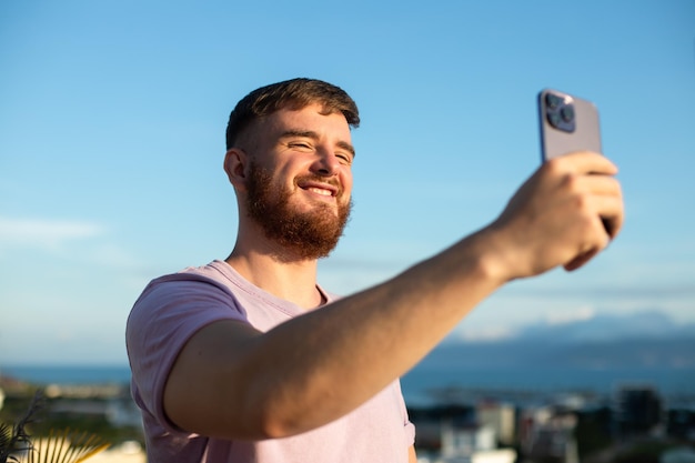 Jeune mec bel homme heureux en mer prend une photo de lui-même selfie à la caméra de son téléphone en utilisant un smartphone pour les médias sociaux à la plage d'été dans un pays exotique tropical Blogger vlog