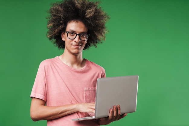 un jeune mec bouclé heureux souriant posant isolé sur un mur vert à l'aide d'un ordinateur portable.