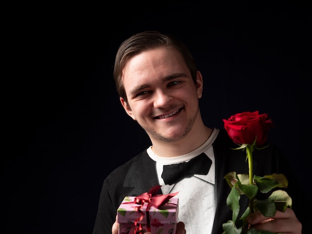 Jeune mec dans un t-shirt noir tenant une rose rouge et un cadeau rose dans ses mains souriant sur fond noir