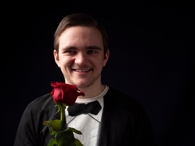 Jeune mec dans un t-shirt noir tient une rose rouge dans ses mains et sourit sur fond noir