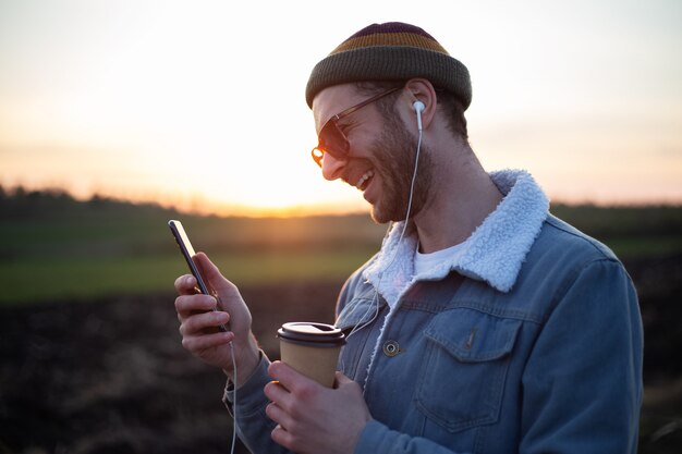 Jeune mec avec des écouteurs prenant des selfies au smartphone avec une tasse de café à la main