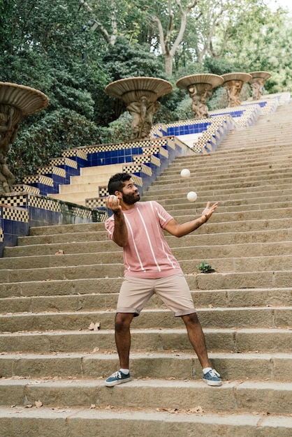 Jeune mec latin jonglant dans les rues ensoleillées de Barcelone Faire un show avec des balles blanches en souriant Vêtu d'un pantalon marron et d'un t-shirt à rayures pastel