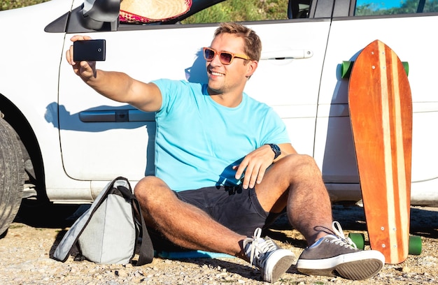 Jeune mec de la mode hipster prenant une photo de selfie cool à côté de sa voiture pendant un voyage sur la route
