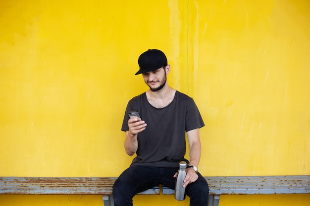 Jeune mec souriant est assis derrière le jaune avec une bouteille en acier à la main et à la recherche dans le smartphone.