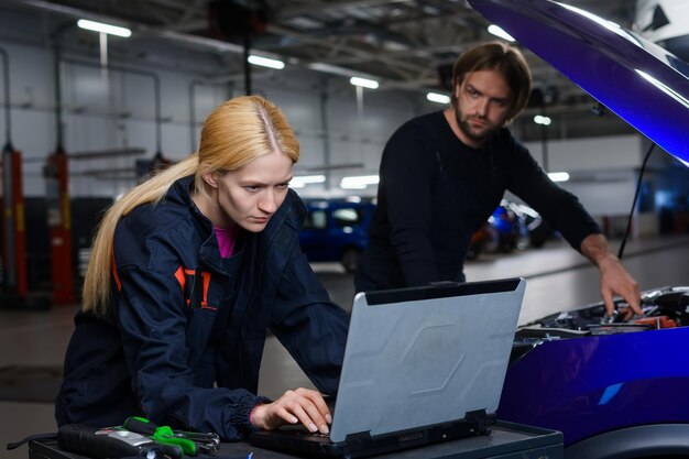 Photo une jeune mécanicienne automobile se tient devant un ordinateur portable de diagnostic et son collègue masculin se tient devant le capot de la voiture diagnostics professionnels à la station-service