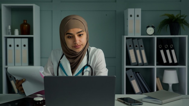 Jeune médecin arabe femme musulmane faisant de la paperasse à la clinique faire de la recherche scientifique utiliser un ordinateur portable