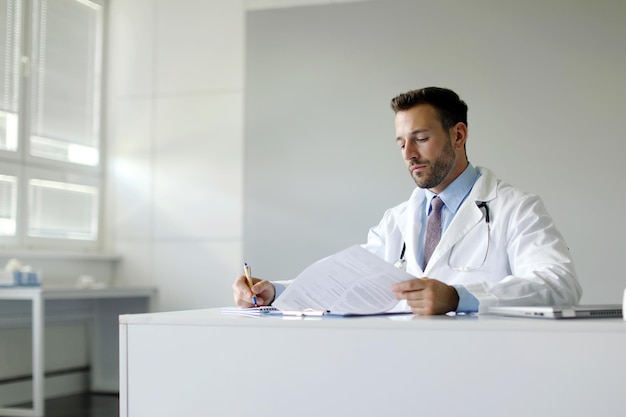 Un jeune médecin assis à une table dans un bureau de clinique écrit et travaille à la vérification