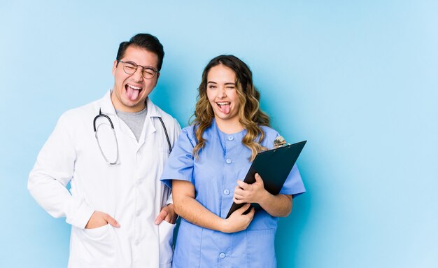 Jeune médecin couple posant dans un mur bleu drôle et sympathique qui sort la langue.