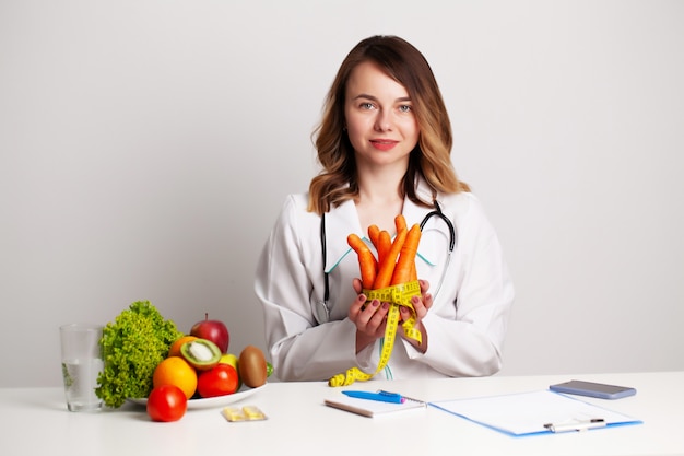 Jeune médecin diététiste à la salle de consultation à la table avec des fruits et légumes frais, travaillant sur un plan de régime