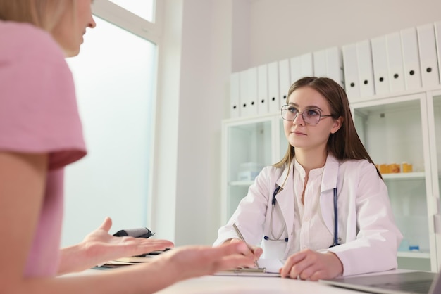 Jeune médecin écoute sa patiente assise à la table de la clinique médicale, une patiente parle