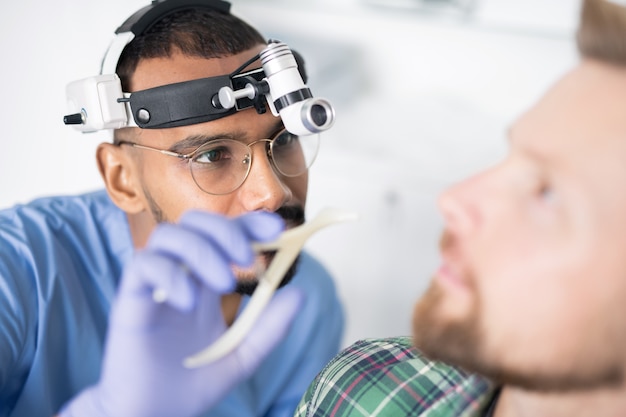 Jeune médecin avec un équipement médical spécial sur sa tête va examiner l'oreille malade du patient avec instrument