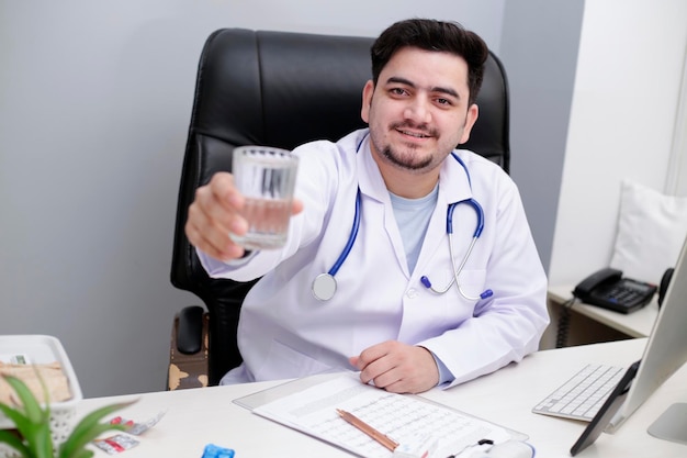 Un jeune médecin est assis sur la chaise à la clinique et tient un verre d'eau à la main