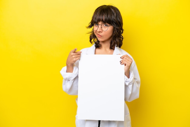 Jeune médecin femme latine isolée sur fond jaune tenant une pancarte vide et pointant vers l'avant