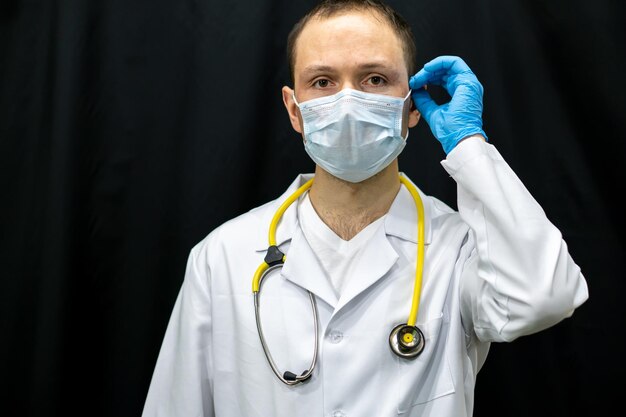 Un jeune médecin en gants bleus et un stéthoscope autour du cou sur un fond noir Portrait d'un médecin avec des instruments médicaux