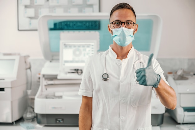 Jeune médecin avec des gants en caoutchouc et un masque facial debout à l'hôpital et montrant les pouces vers le haut. Le virus Corona est en baisse.