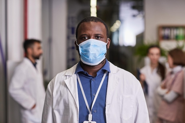 Jeune médecin généraliste afro-américain en blouse de laboratoire et masque debout devant la caméra contre ses collègues à l'hôpital