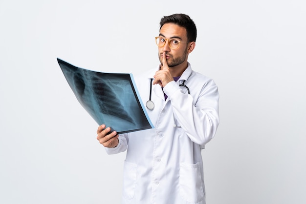 Jeune médecin homme tenant une radiographie isolée sur un mur blanc faisant un geste de silence