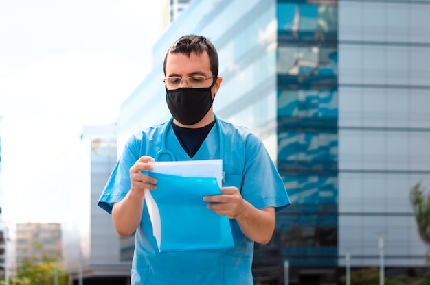 Jeune médecin latin, portant des lunettes et un manteau bleu, à l'extérieur de l'hôpital, portant un embout buccal pour se protéger du coronavirus, lisant un historique médical.