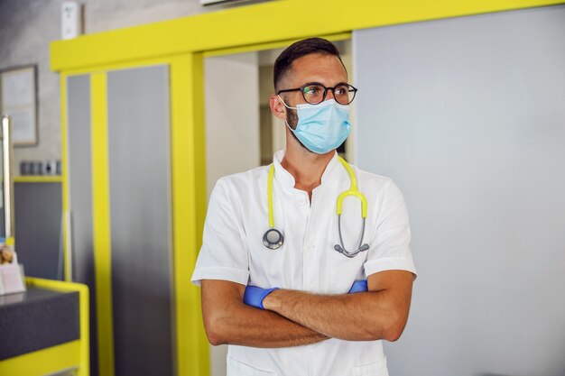 Jeune médecin avec masque facial, gants en caoutchouc et en uniforme blanc stérile debout à l'hôpital avec les bras croisés et regardant ailleurs.